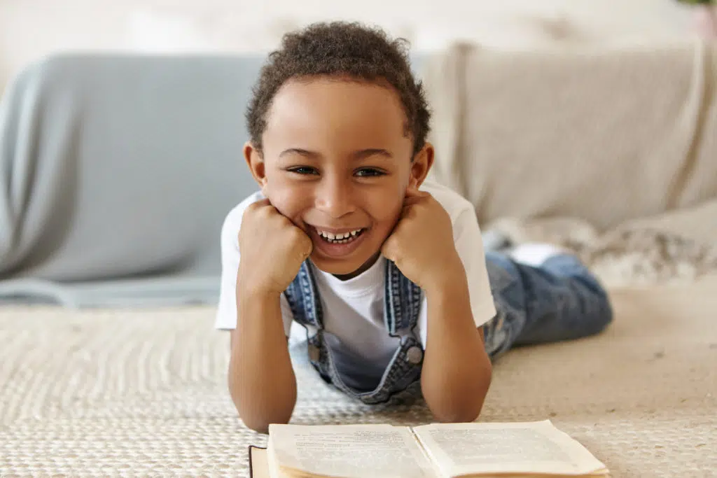 Aide aux devoirs : un enfant allongé sur le tapis et qui lit un livre avec le sourire 